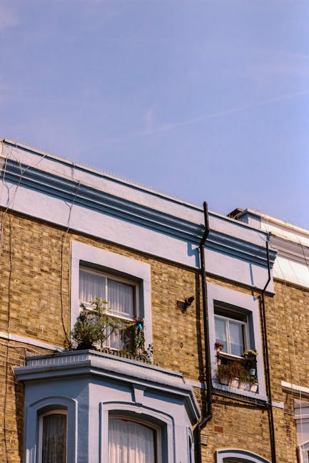 A tall, multi-storey residential building with a brick exterior and white window frames. On the second floor, there is a bay window with a small ledge decorated with potted plants and flowers, and the window has a white curtain partially visible inside. The building has visible external black drainpipes running vertically along the facade and a white, flat roof with some piping and a vent system attached. Above, the sky is clear and blue with a few wispy clouds. This scene is typical of urban house architecture and may be part of a property involved in home relocation or furniture transport services, as provided by Movers Finsbury Park. The image captures the exterior environment where moving logistics, such as loading or unloading at the door or nearby side areas, could take place, highlighting the context of house removals in Finsbury Park, N4.