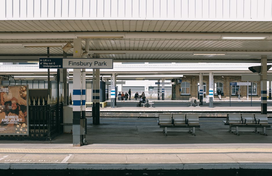 The image shows Finsbury Park railway station platform with a white sign displaying the station name. The platform is sheltered by a metal roof with horizontal paneling, and several metal benches are positioned along the waiting area. In the background, multiple tracks can be seen extending beneath the shelter, with a few passengers sitting and standing, some waiting for trains. A poster advertising 'Romeo and Juliet' is visible on the left side near a black metal fence, which separates the platform from the station entrance. The environment is well-lit with natural daylight, and the scene depicts a typical setting for house removals and furniture transport, where careful planning is required when accessing the station for home relocation services, as sometimes tight access areas are involved. Movers Finsbury Park often assist clients with logistics involving loading and unloading of furniture and boxes, with attention to safety and efficiency in busy or constrained spaces, especially during packing and moving operations.