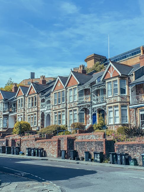 A row of Victorian-style terraced houses situated along a residential street, featuring ornate cast-iron balconies, bay windows, and gabled roofs with decorative trim. The houses are constructed with 