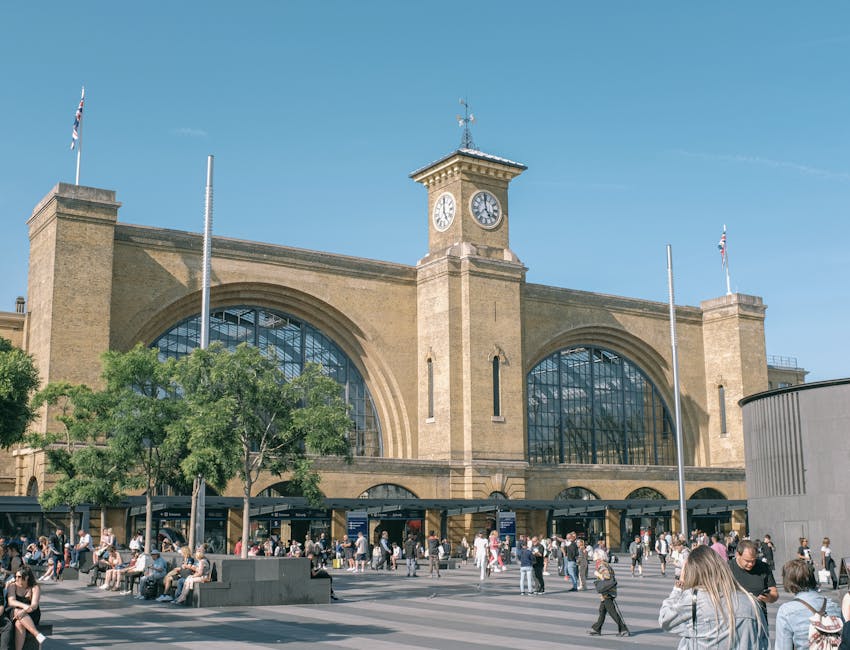 The exterior of a historic train station building with large arched windows and a prominent clock tower at the centre, under a clear blue sky. The station’s stone facade is visible with flags mounted on flagpoles along the front. In the foreground, a busy pavement area is populated with numerous pedestrians, some sitting on benches and others walking. There are trees providing partial shade near the entrance, and the area appears lively with people waiting, chatting, or passing by. Although no moving or packing activities are depicted, the setting suggests a location relevant to home relocation or transport services, aligned with the context of house removals and furniture transport by Movers Finsbury Park, capturing a typical scene associated with transit hubs used in moving logistics.