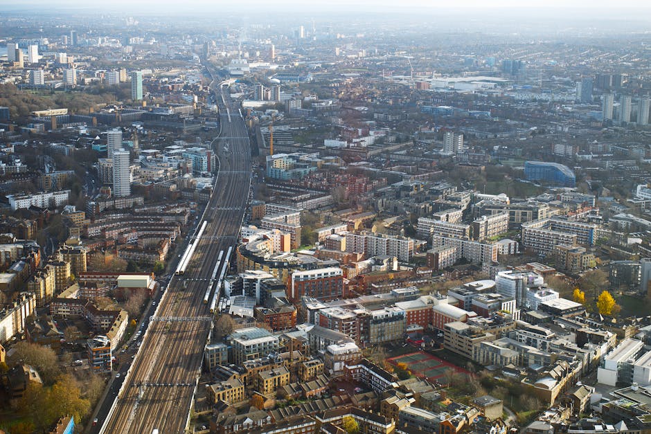 Aerial view of a cityscape with a multilane railway line running through the urban area, surrounded by numerous mid-rise and high-rise buildings, residential complexes, commercial structures, and tree-lined streets. The railway tracks are visibly elevated in some sections, with a train approaching or departing in the distance. The environment appears to be daytime with natural lighting, showing a mix of modern and older architecture. This scene is indicative of a busy metropolitan area, where moving and relocation services by Movers Finsbury Park may frequently operate, particularly in densely populated districts near Finsbury Park station. The image captures the expansive urban setting suitable for house removals, furniture transport, and packing and moving logistics within the city's residential and commercial zones.
