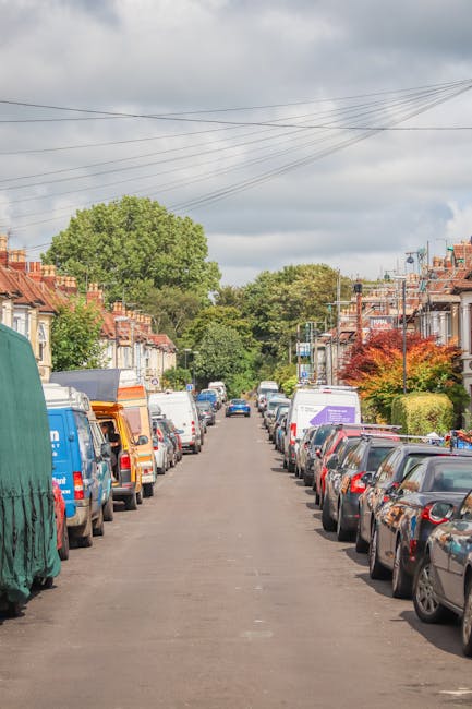 A residential street with rows of terraced houses featuring red brick facades and small front gardens, lined with parked cars on both sides, including sedans, hatchbacks, and vans. Some vans are covered with protective blankets and plastic wrap, indicating they are part of a house removal process. A moving trolley and loading straps are visible near the vehicles, suggesting active loading or unloading of furniture and boxes. The street is paved with asphalt and has overhead power lines, with lush green trees and shrubs adding natural elements to the scene. The sky above is cloudy, and the environment appears calm and quiet, typical of a neighborhood preparing for a household relocation. This scene exemplifies the logistics involved in furniture transport and packing during a home removal, with a focus on safe vehicle loading and organized street parking, as carried out by Movers Finsbury Park.