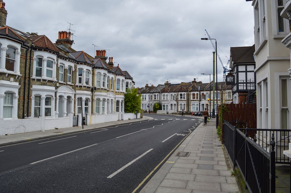 A quiet residential street on a cloudy day featuring Victorian-style terraced houses with white facades, bay windows, and red tiled roofs. The pavement runs along the right side of the image, bordered by black metal railings and small front gardens. Several cardboard boxes and packaged belongings are visible on the pavement near the entrance of a property, indicating preparations for home relocation. An orange and black moving blanket, along with a trolley, are seen being used in the loading process. The street is lined with street lamps, parked cars, and a large construction crane visible in the background, suggesting ongoing development. The overall scene depicts an urban environment where furniture transport and packing are underway, with the presence of a moving company, Movers Finsbury Park, supporting the logistics of house removals on Stroud Green Road.
