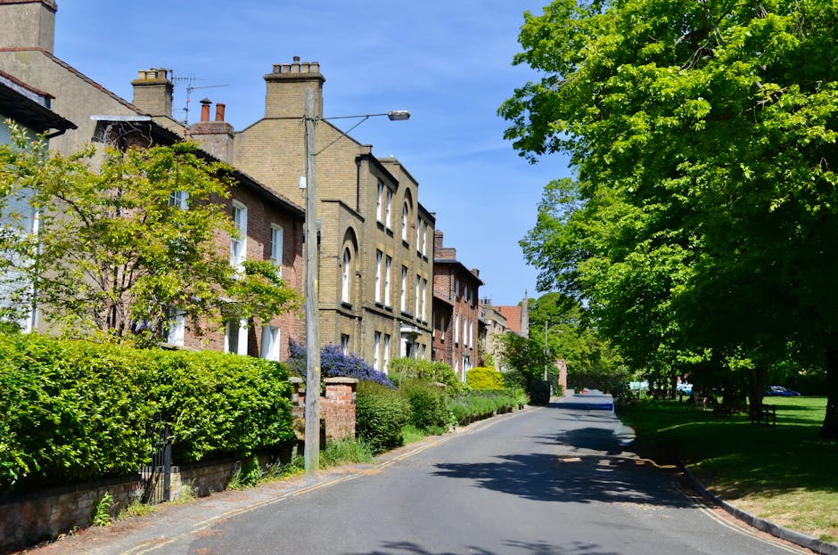 A residential street with rows of terraced houses featuring red brick facades and small front gardens, lined with parked cars on both sides, including sedans, hatchbacks, and vans. Some vans are covered with protective blankets and plastic wrap, indicating they are part of a house removal process. A moving trolley and loading straps are visible near the vehicles, suggesting active loading or unloading of furniture and boxes. The street is paved with asphalt and has overhead power lines, with lush green trees and shrubs adding natural elements to the scene. The sky above is cloudy, and the environment appears calm and quiet, typical of a neighborhood preparing for a household relocation. This scene exemplifies the logistics involved in furniture transport and packing during a home removal, with a focus on safe vehicle loading and organized street parking, as carried out by Movers Finsbury Park.